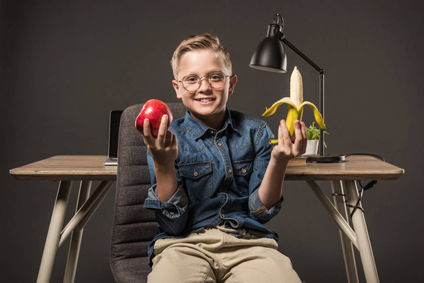 Lächelnder kleiner Junge in Brille mit Apfel und Banane am Tisch mit Lampe, Pflanze und Laptop auf grauem Hintergrund  - Foto, Bild