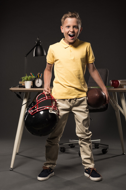 excited screaming schoolboy holding american football helmet with ball and standing near table with books, plant, lamp, colour pencils, apple, clock and textbook on grey background  - Photo, Image