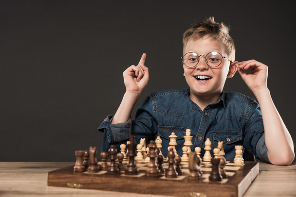 glücklicher kleiner Junge mit Brille macht Idee Geste mit dem Finger am Tisch mit Schachbrett isoliert auf grauem Hintergrund  - Foto, Bild