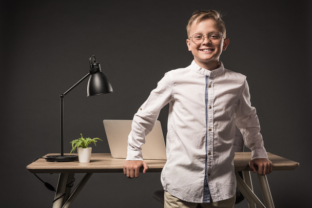 smiling little boy in eyeglasses standing near table with plant, laptop and plant on grey background  - Photo, Image