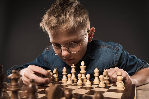fokussierter kleiner Junge mit Brille spielt Schach am Tisch isoliert auf grauem Hintergrund  - Foto, Bild