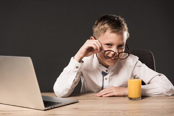 smiling little boy putting on eyeglasses at table with laptop and glass of juice on grey background  - Foto, immagini