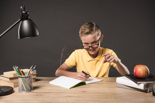 happy schoolboy in eyeglass pointing by finger and doing homework at table with lamp, books, colour pencils and textbook on grey background  - Photo, Image