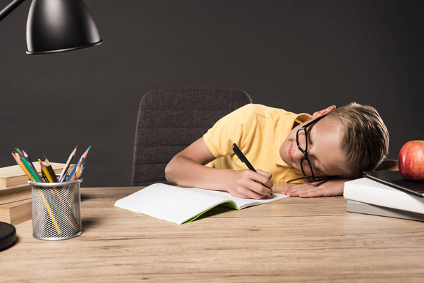 Schüler in Brille legt sich auf den Tisch und erledigt Hausaufgaben am Tisch mit Lampe, Büchern, Farbstiften und Lehrbuch auf grauem Hintergrund  - Foto, Bild