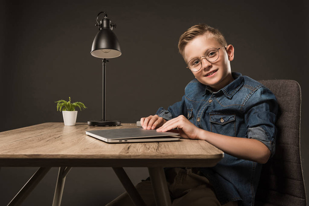 smiling little boy in eyeglasses opening laptop at table with plant and lamp on grey background  - Photo, Image