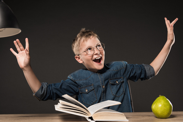 excited school boy in eyeglasses gesturing by hands while reading book at table with lamp and pear on grey background  - Photo, Image
