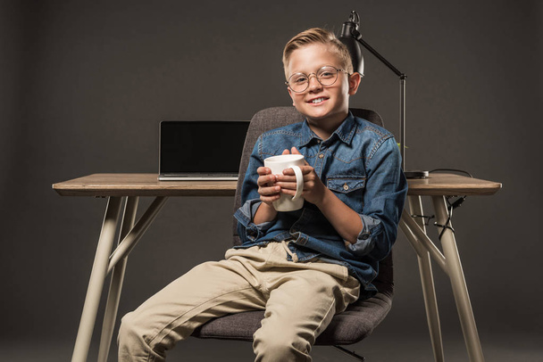 smiling little boy in eyeglasses sitting on chair with coffee cup near table with laptop and lamp on grey background  - Zdjęcie, obraz