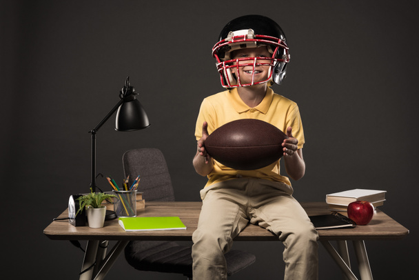smiling schoolboy in american football helmet holding ball and sitting on table with books, plant, lamp, colour pencils, apple, clock and textbook on grey background  - Foto, Bild