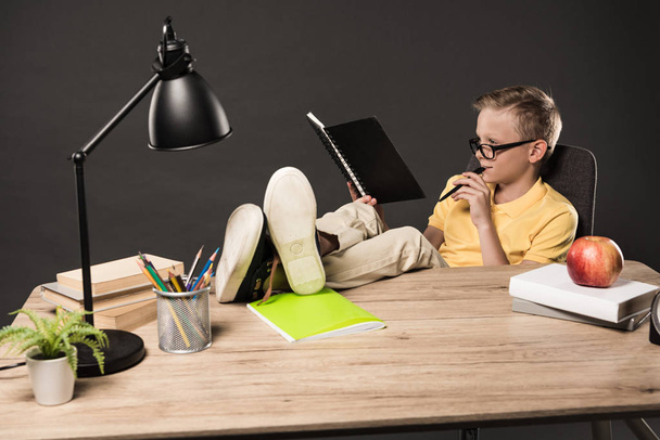schoolboy in eyeglasses doing homework with legs on table with books, plant, lamp, colour pencils, apple, clock and textbook on grey background  - Photo, Image