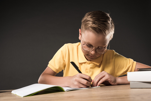 fokussierter Schuljunge mit Brille, der im Lehrbuch am Tisch mit einem Stapel Bücher auf grauem Hintergrund schreibt  - Foto, Bild