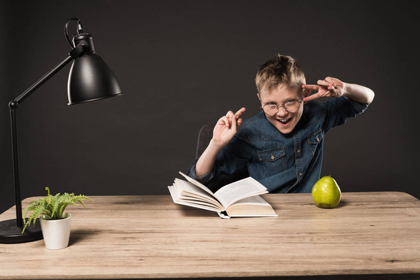 excited little boy in eyeglasses doing peace signs at table with book, pear, plant and lamp on grey background  - Foto, immagini