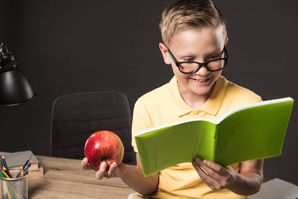 smiling schoolboy in eyeglasses holding apple and doing homework near table with colour pencils, lamp and books on grey background  - Foto, immagini