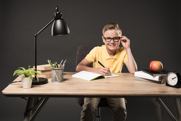 маленький школьник в очках делает домашнюю работу за столом с книгами, растениями, лампами, цветными карандашами, яблоками, часами и учебниками на сером фоне
  - Фото, изображение