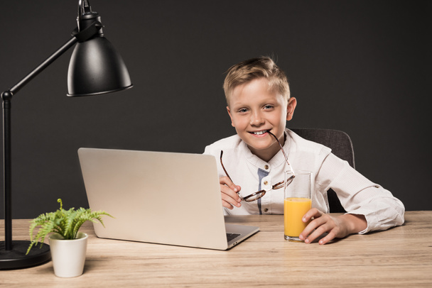 usmívající se malý chlapec drží brýle a sedí u stolu s sklenici šťávy, laptop, rostlin a lampu na šedém pozadí  - Fotografie, Obrázek