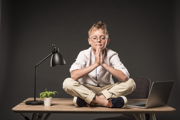 little boy with closed eyes in eyeglasses meditating and sitting on table with laptop, plant and lamp on grey background  - Photo, Image