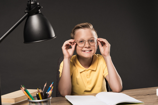 smiling schoolboy adjusting eyeglasses and sitting at table with lamp, colour pencils, books and textbook on grey background  - Photo, Image