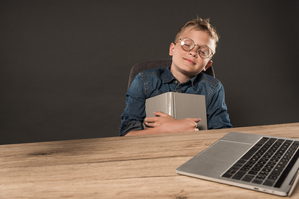 little schoolboy in eyeglasses sleeping with book at table with laptop on grey background  - Foto, Imagem