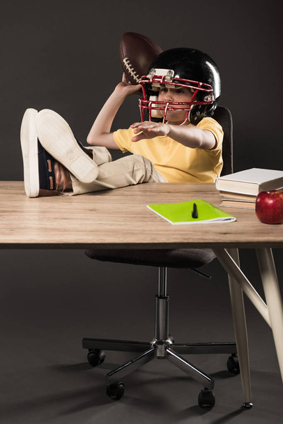 scolaretto sorridente in casco da calcio americano con palla in mano e seduto con le gambe sul tavolo con libri, mela e libro di testo su sfondo grigio
  - Foto, immagini