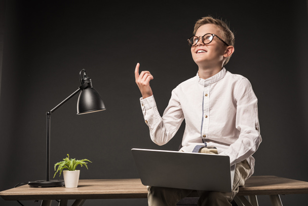 uśmiechający się mały chłopiec w okularach robi pomysł gest palcem i przy użyciu laptopa siedząc na stole z lampa i roślin na szarym tle  - Zdjęcie, obraz