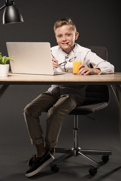 lächelnder kleiner Junge mit Brille und am Tisch sitzend mit Laptop, Saftglas, Pflanze und Lampe auf grauem Hintergrund  - Foto, Bild