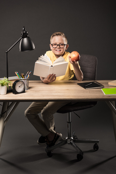 smiling schoolboy in eyeglasses holding apple and reading book at table with books, plant, lamp, colour pencils, clock and textbook on grey background  - Foto, immagini