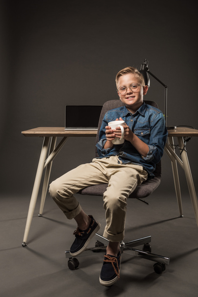 little boy in eyeglasses holding coffee cup and sitting on chair near table with laptop and lamp on grey background  - Photo, Image