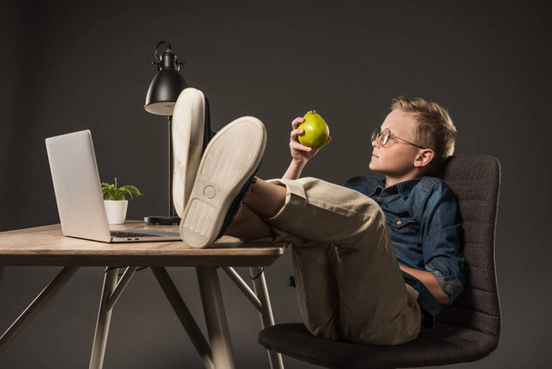 vista lateral do menino em óculos segurando pêra e sentado com as pernas na mesa com laptop, planta e lâmpada no fundo cinza
  - Foto, Imagem
