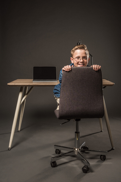 smiling little boy in eyeglasses sitting on chair near table with laptop and lamp on grey background  - Foto, imagen