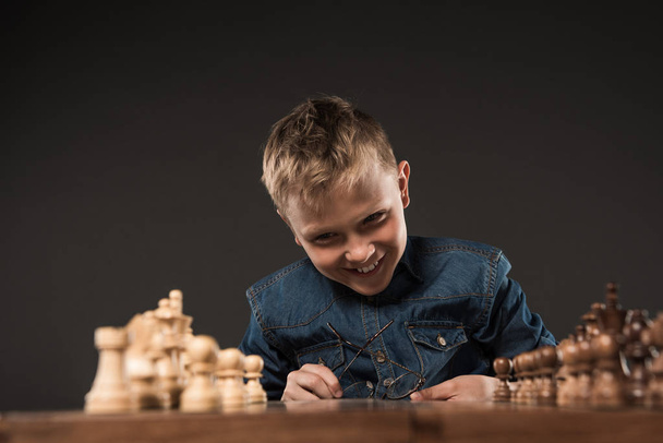 Gelukkig jongetje brillen houden en kijken naar schaakbord aan tafel geïsoleerd op een grijze achtergrond  - Foto, afbeelding