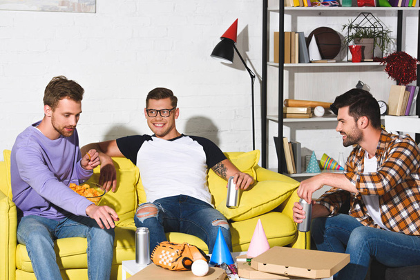 knappe glimlachend jonge mannen bier drinken en eten snacks thuis partij  - Foto, afbeelding