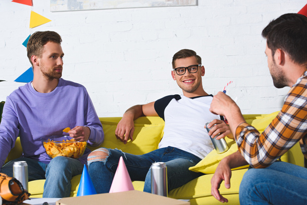 smiling young male friends drinking beer and eating snacks at home party  - Photo, image