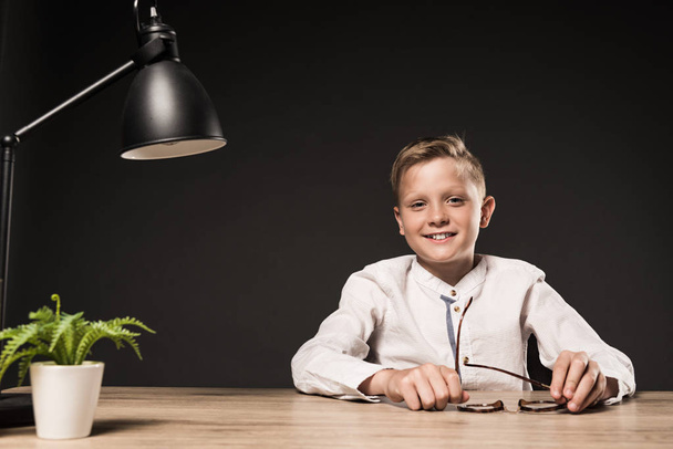 happy little boy sitting at table with eyeglasses, plant and lamp on grey background  - Valokuva, kuva