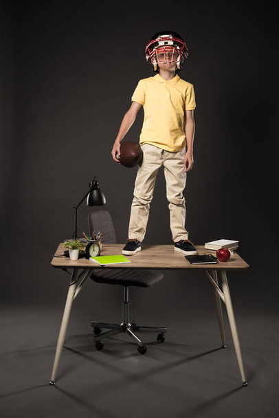 schoolboy in american football helmet holding ball and standing on table with books, plant, lamp, colour pencils, apple, clock and textbook on grey background  - Foto, Bild