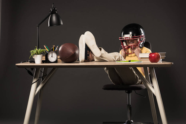 colegial sonriente en casco de fútbol americano sentado con las piernas en la mesa con bola, libros, planta, lámpara, lápices de color, manzana, reloj y libro de texto sobre fondo gris
  - Foto, imagen