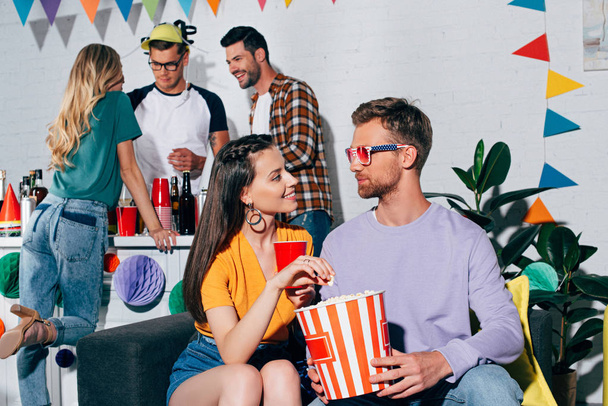young man and woman looking at each other while drinking beer and eating popcorn at home party - Foto, Bild