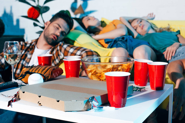 lähikuva pizzalaatikosta, muovikupeista ja baseball-laitteista pöydällä ja humalaiset nuoret nukkuvat takana
 - Valokuva, kuva