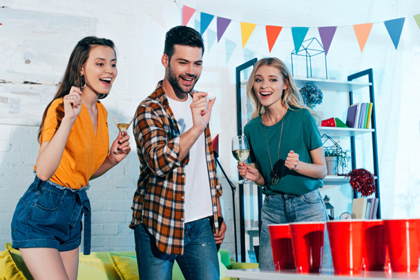 smiling girls with glasses of wine looking at young man playing beer pong at home party  - Foto, Imagen