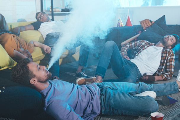 young man smoking electronic cigarette while drunk friends sleeping after party  - Photo, Image