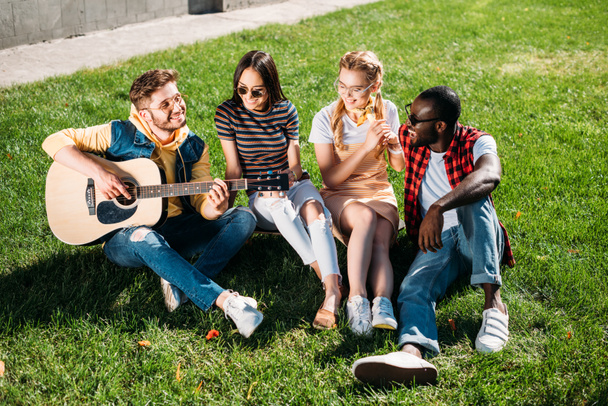 interracial group of friends with acoustic guitar resting on green grass in park on summer day - Foto, Bild