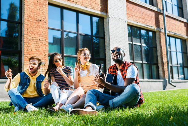 interracial group of happy friends with acoustic guitar resting on green grass on summer day - 写真・画像