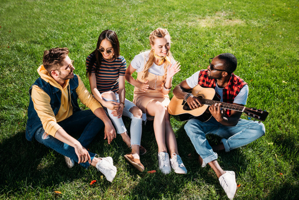 interracial group of friends with acoustic guitar resting on green grass in park on summer day - Fotó, kép