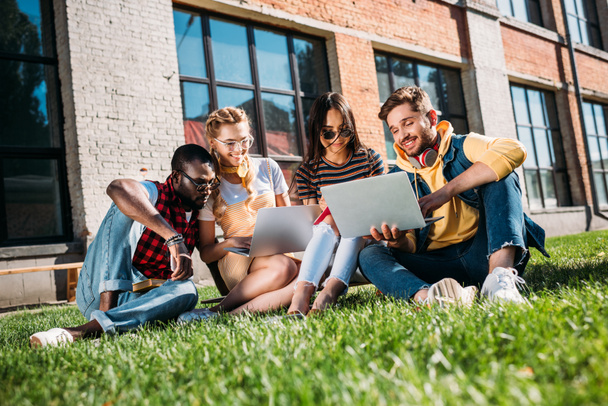 interracial group of students with laptops and notebooks studying on green lawn together - Фото, изображение