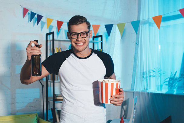 smiling young man holding popcorn box and beer bottle at home party - Foto, imagen