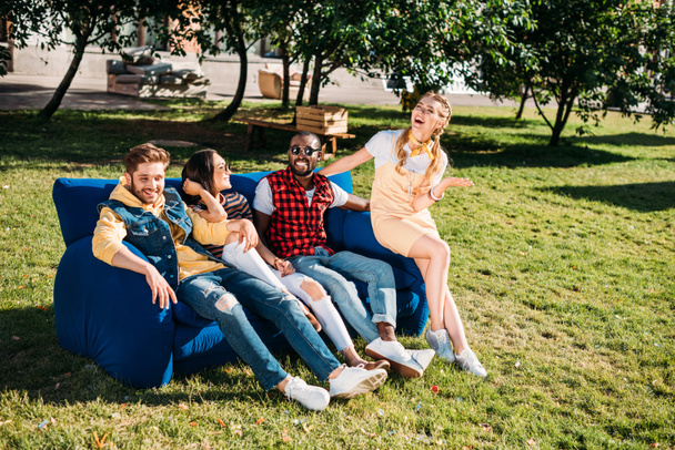 lächelnde interrassische Freunde, die gemeinsam auf blauem Sofa im Park liegen - Foto, Bild
