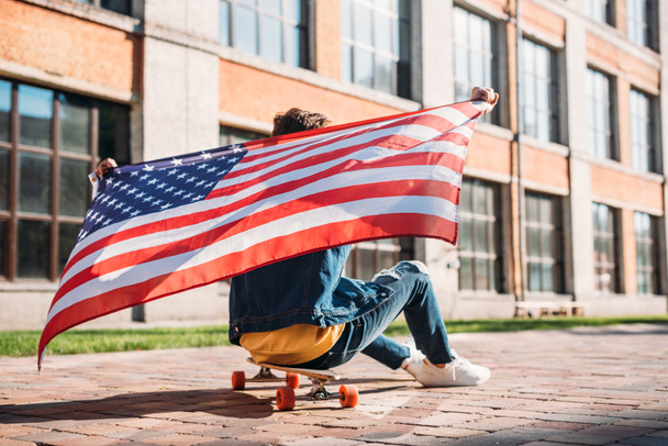 back view of man with american flag sitting on longboard on street - Foto, imagen