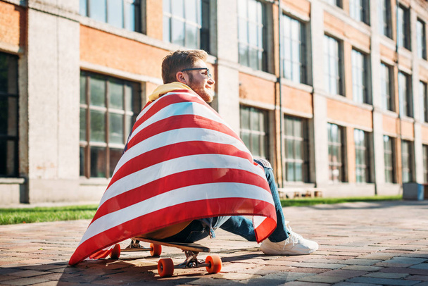 back view of young man with american flag sitting on longboard on street - Foto, immagini