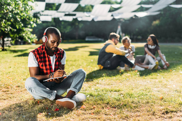 enfoque selectivo del hombre afroamericano en auriculares con portátil descansando sobre hierba verde con amigos multirraciales con ordenador portátil detrás
 - Foto, imagen