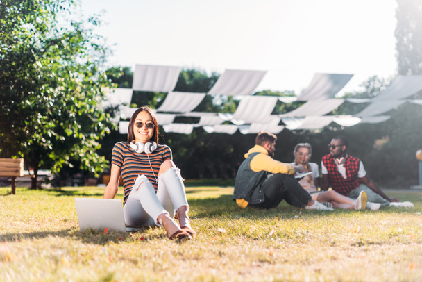 selective focus of young asian woman with laptop and multiracial friends behind resting on green grass in park - Fotoğraf, Görsel