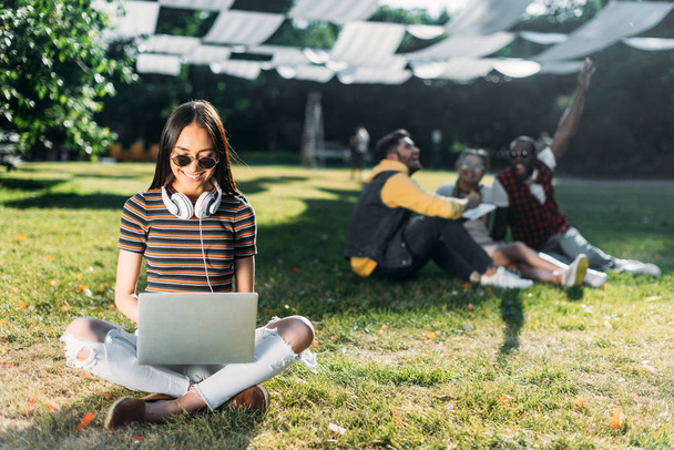 selective focus of smiling asian woman using laptop and multiracial friends behind resting on green grass in park - Foto, Bild