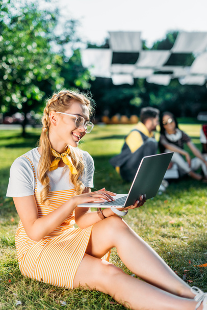 selectieve aandacht van jonge lachende vrouw met laptop en vrienden achter rustend op groene gras in park - Foto, afbeelding